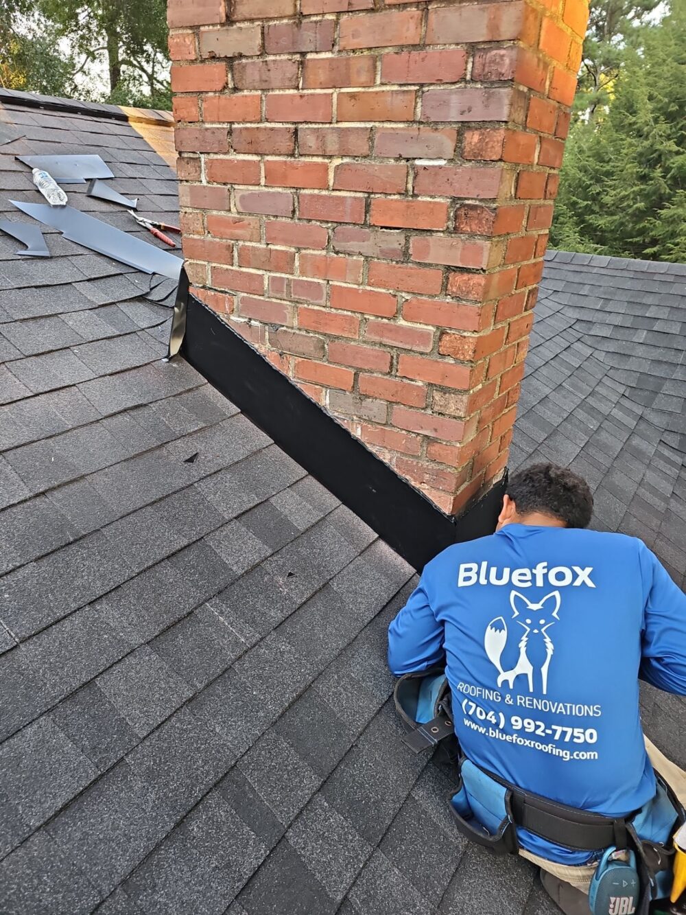 Roofing installer in Blue Fox Roofing shirt adds finishing touches to the chimney flashing on the roof.
